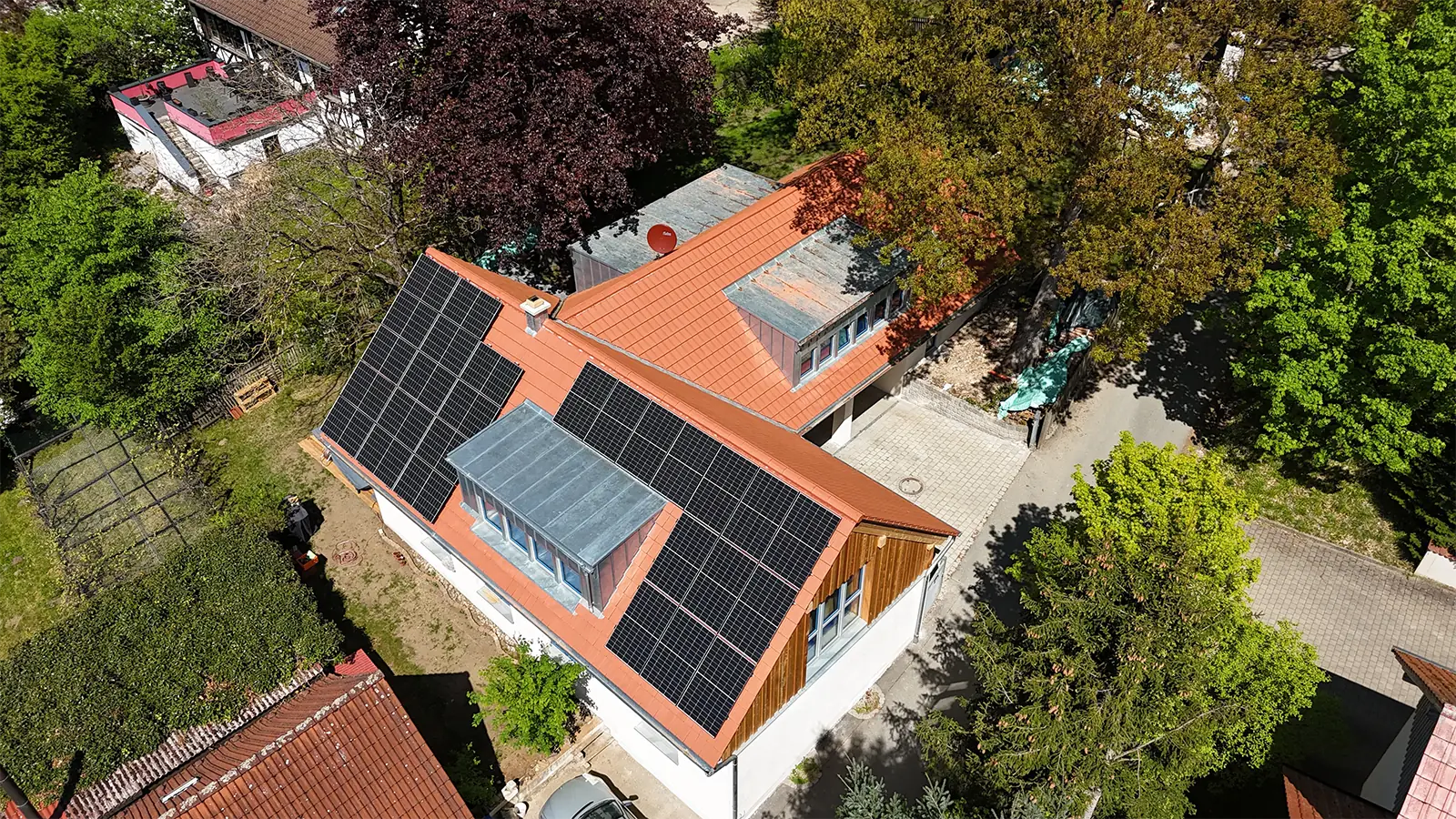 Vogelperspektive des sanierten Hauses. Auf dem Dach mit roten Ziegeln sind flächendeckend Solarzellen angebracht. Um das Gebäude sind die Zufahrt, Gärten und viele Bäume zu erkennen.