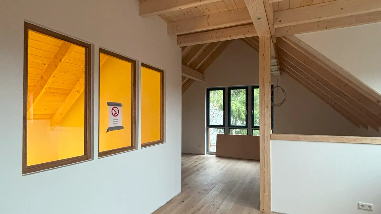 Innenansicht des sanierten Dachgeschosses. Die Räumlichkeiten sind offen, in einen abgetrennten Raum kann man durch drei innenliegende Fenster blicken. Im abgetrennten Raum leuchtet ein gelbes Licht.