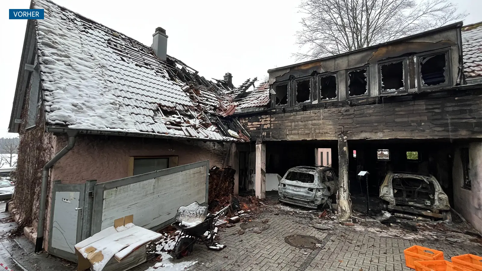 Vorheransicht des Hauses nach Brandschaden. Die Garage ist komplett ausgebrannt, die Fenster sind geborsten, die Fassade schwarz vor Ruß. In der Garage stehen zwei ausgebrannte Fahrzeuge. Das Dach ist teilweise eingestürzt.