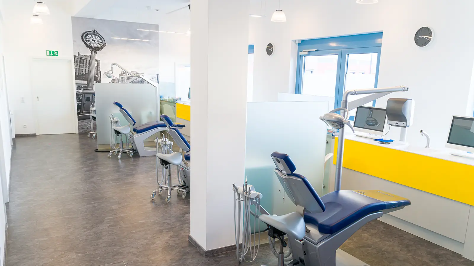 Fertig sanierte Praxisräume mit mehreren Behandlungssektionen, die durch undurchsichtige Glaswände getrennt sind. Der Raum ist durch Deckenleuchten und große Fenster hell erleucht. Im Vordergrund sind die Behandlungsstühle und -lichter zu sehen.
