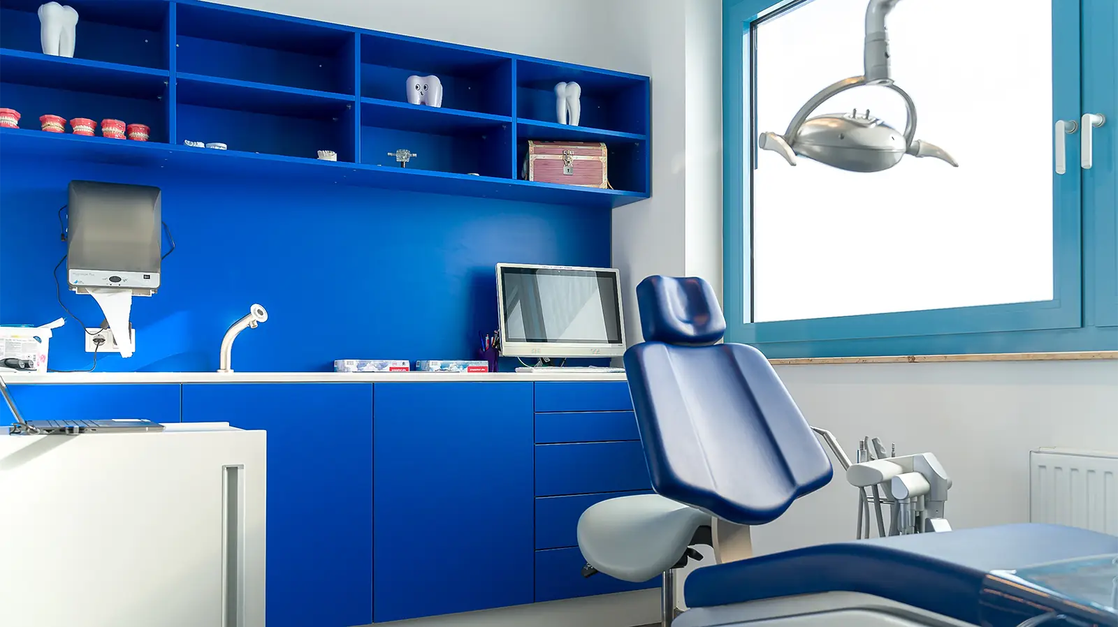 Behandlungszimmer nach den abgeschlossenen Sanierungsarbeiten. Im Vordergrund ist ein blauer Behandlungsstuhl, daneben eine einstellbare Leuchte. Im Hintergrund sind leuchtend blaue Schränke, ein Computer und Sanitäranlagen zu sehen.