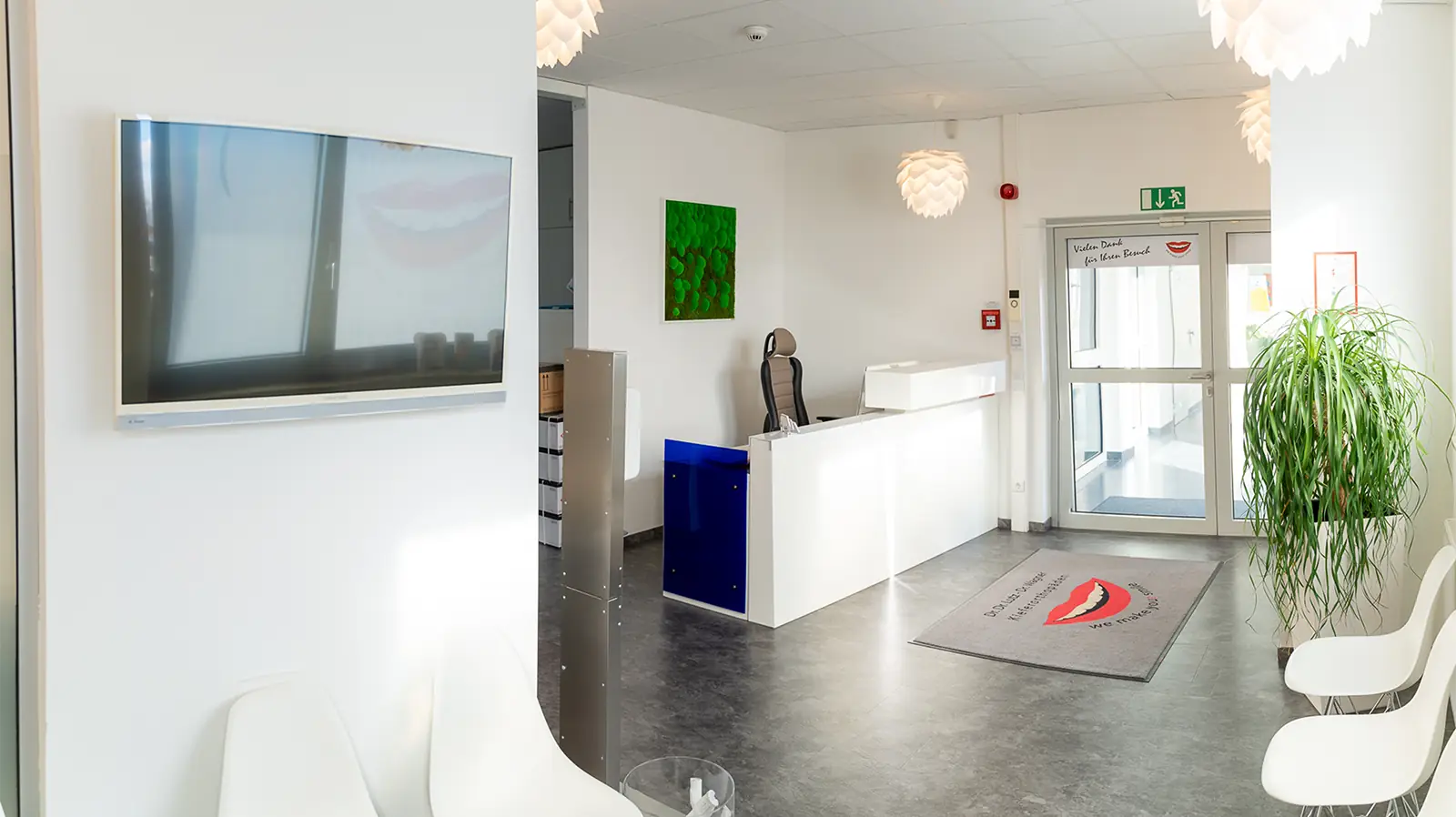 Empfangsbereich der Praxis nach den vollendeten Sanierungsarbeiten. Im Vordergrund ist eine Pflanze zu sehen, auf der rechten Seite ist der Wartebereich mit drei weißen Stühlen zu sehen. Im linken Bereich ist der Empfang.