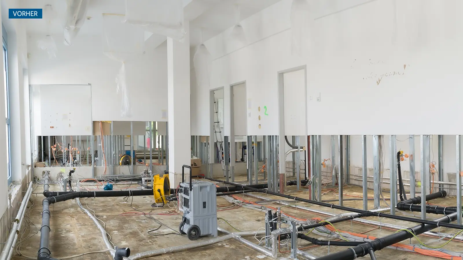 Blick auf einen unfertigen Raum mit freiliegenden Rohren und Kabeln auf dem Boden. An den Wänden sind Metallrahmen für Trockenbau sichtbar. Einige Bereiche sind mit Plastikfolie abgedeckt.