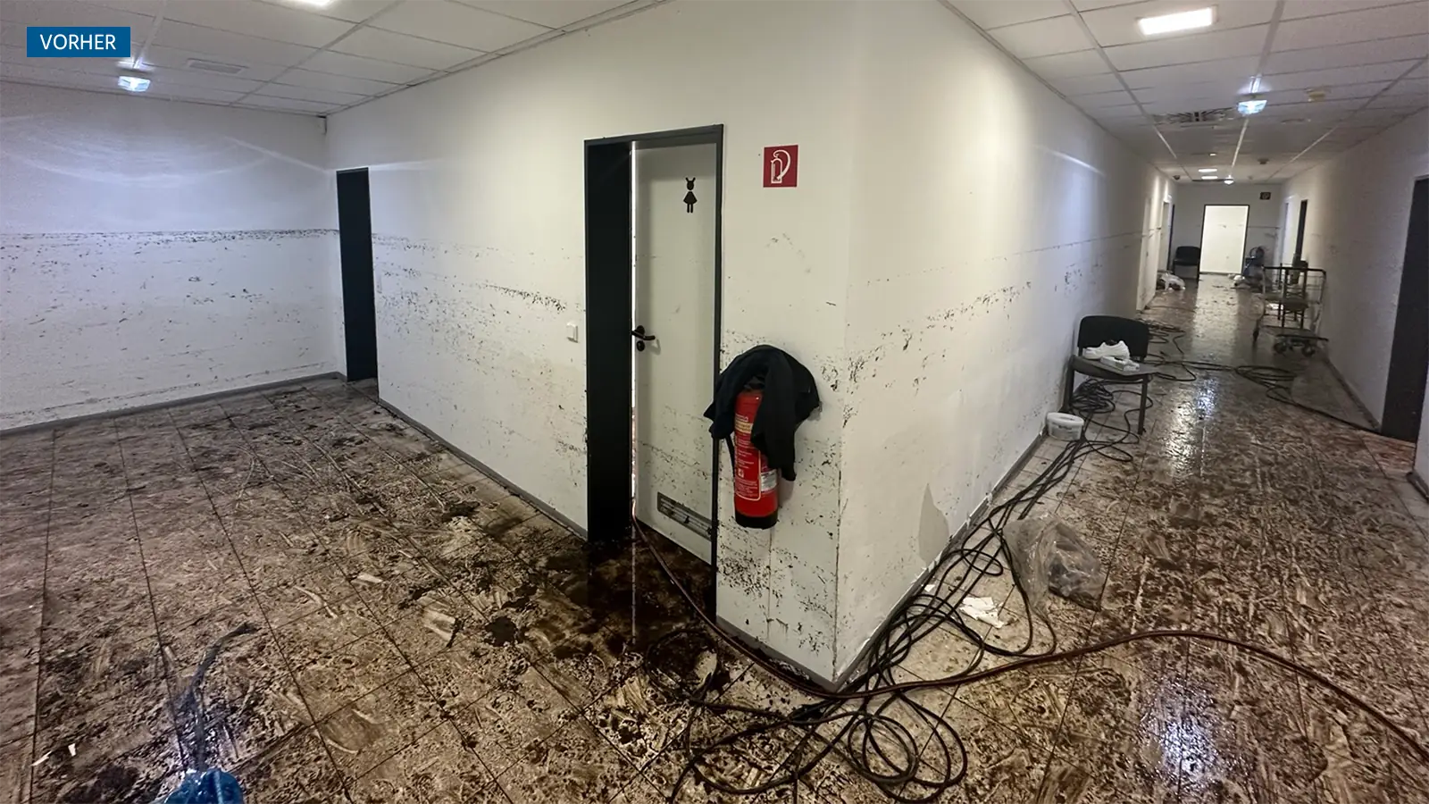 Der Flur des Mitarbeiterbereichs nach der Überflutung. Die Schäden durch die Niederschlagsmengen sind an der Wand zu sehen, der Boden ist schmutzig durch die Überflutung. Über den Boden verteilt liegen noch Kabel und Schläuche zum Abpumpen des Wassers.