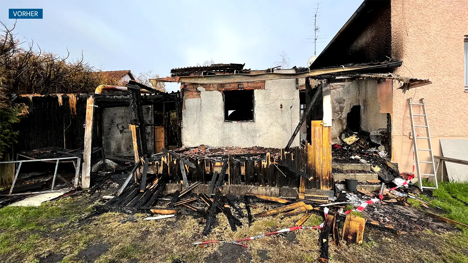 Die Garage und Teile des Wohnhauses nach dem Garagenbrand. Von der Garage bleibt fast nichts übrig. Im vorderen Teil sieht man kleine Reste des verbrannten Holzes, nur noch die Rückwand steht. Am Wohnhaus sind starke Brandspuren zu sehen.