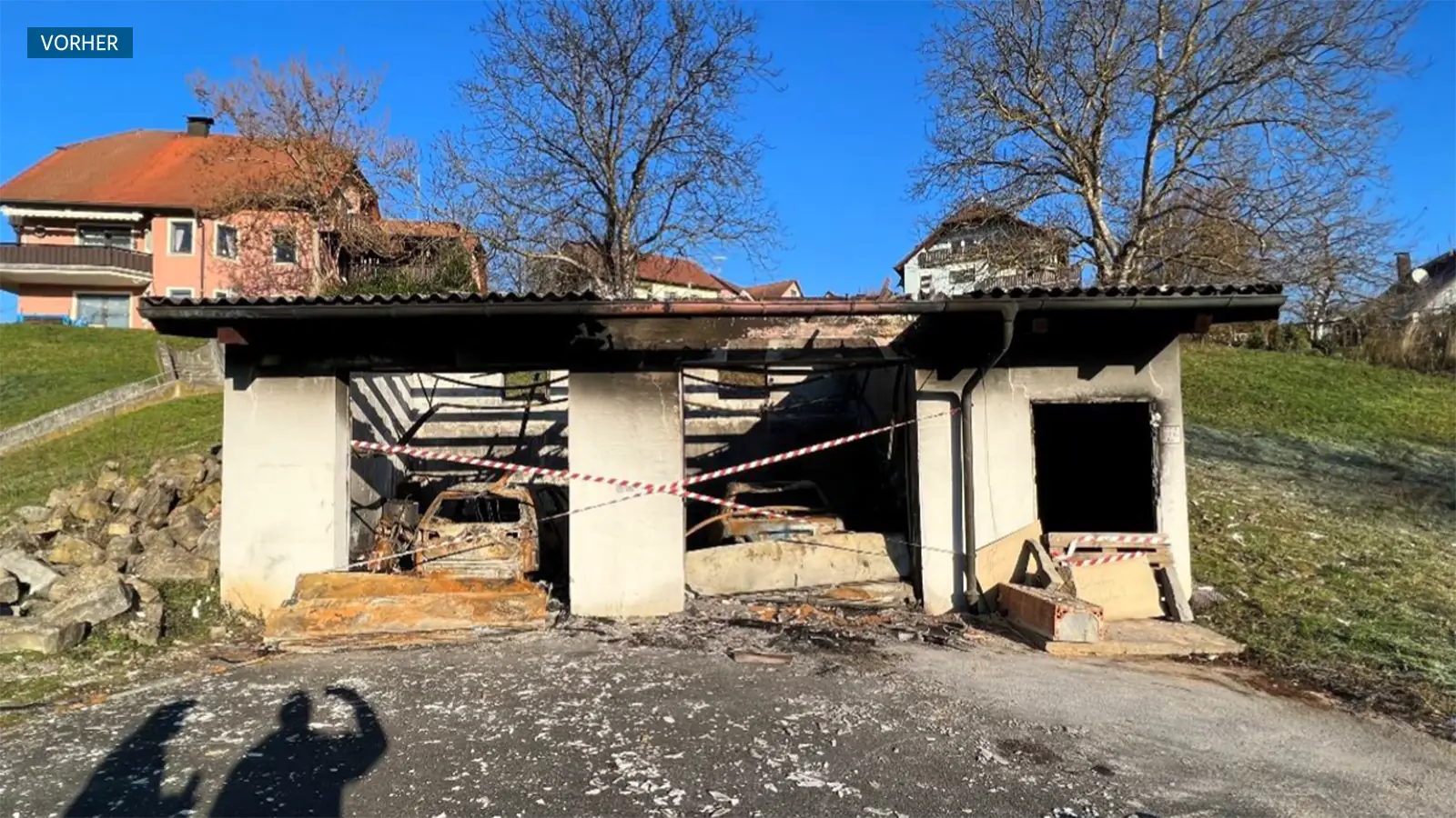 Das Bild zeigt die Doppelgarage nach dem Fahrzeugbrand. Die Sonne wirft Schatten der Holzbalken durch das kaputte Dach. Hinter den kollabierten Toren sieht man die ausgebrannten Fahrzeuge. Die Garage ist mit Absperrband abgeriegelt.