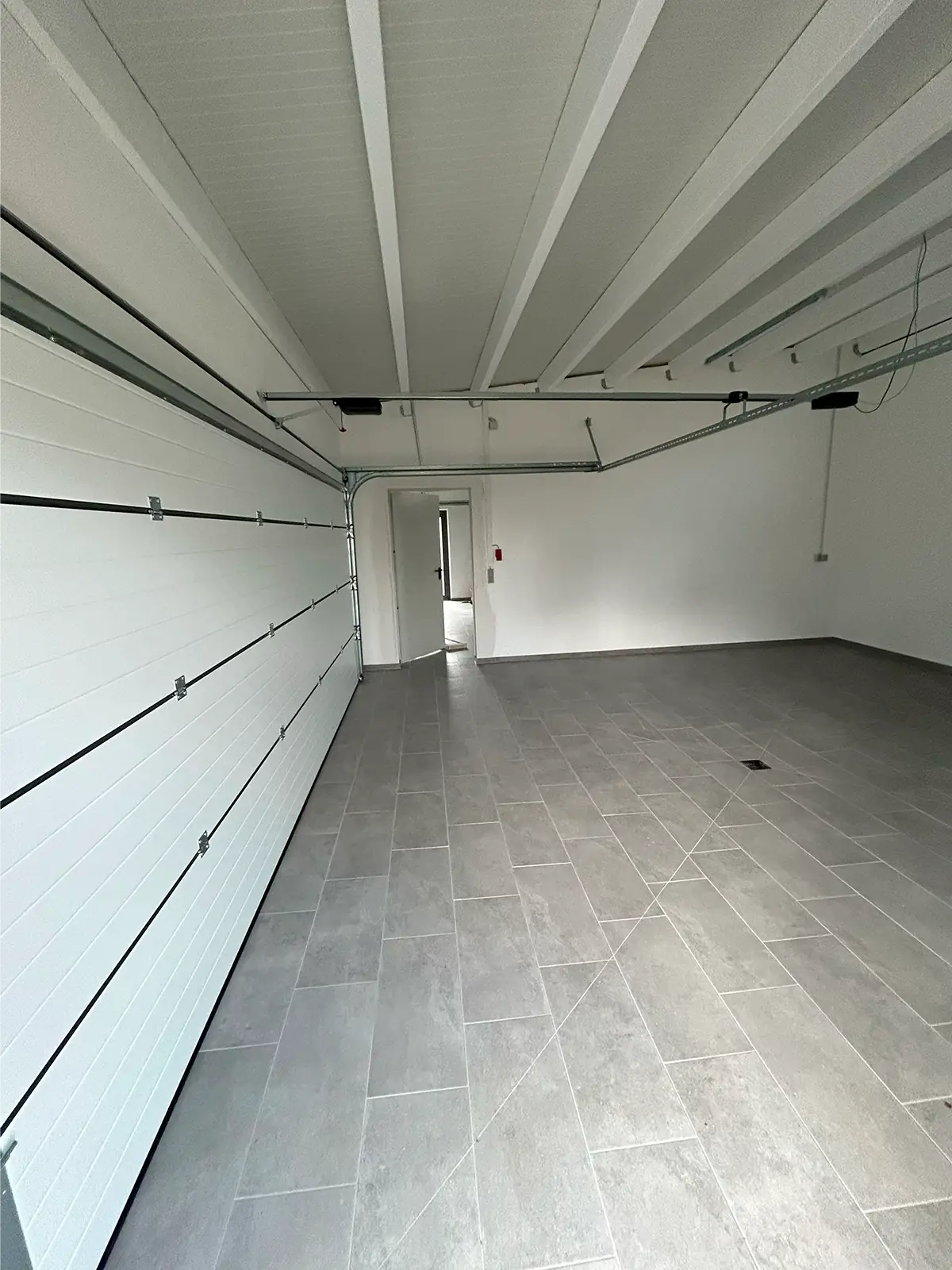 Das Bild zeigt die sanierte Doppelgarage von Innen. Die Wände sind weiß gestrichen, der Boden ist grau gefließt. Links ist das geschlossene Garagentor zu sehen. Im Hintergrund führt eine offene Tür zum nächsten Teil der Doppelgarage.