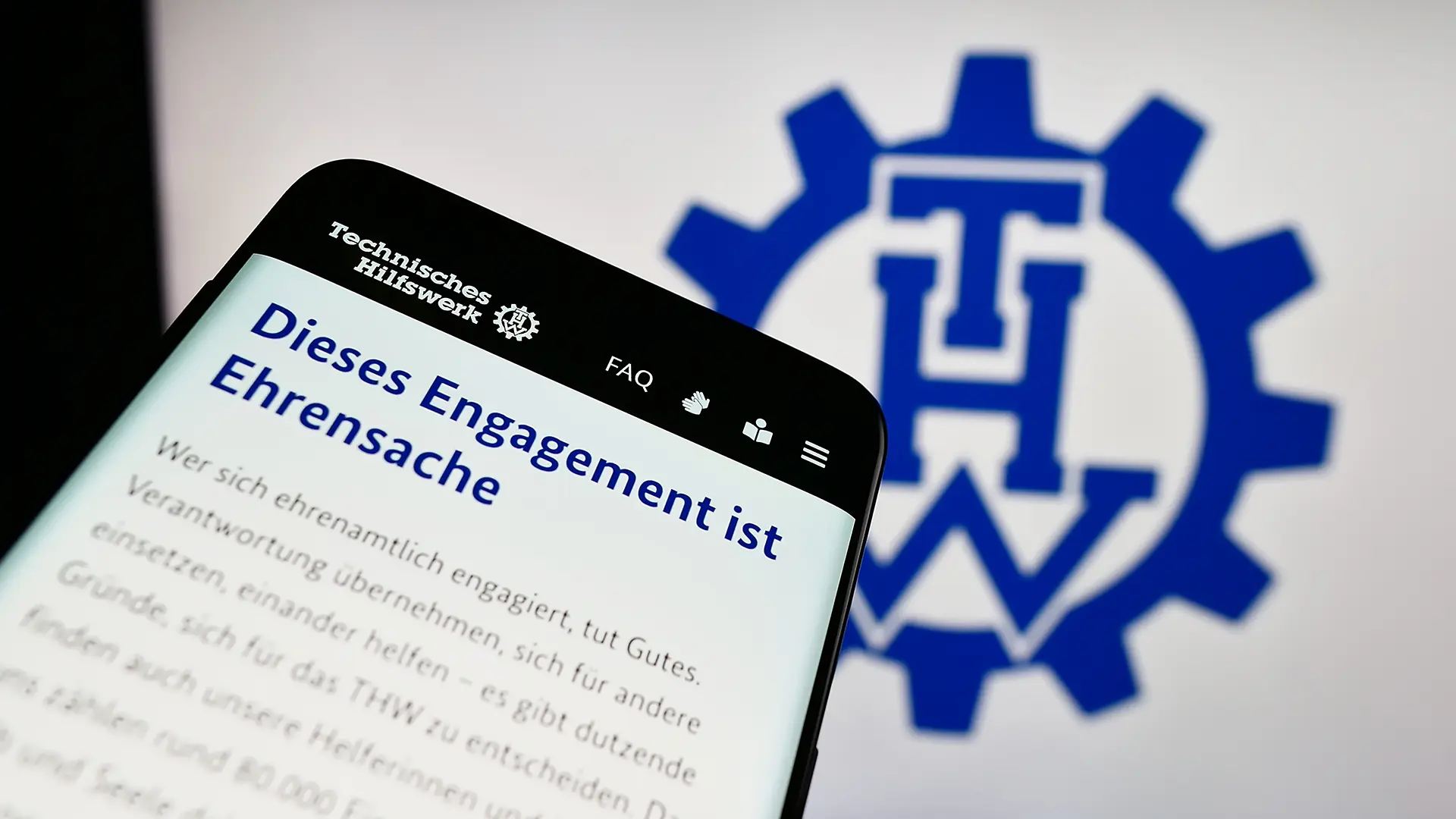 Ein Smartphone vor dem Hintergrund des THW Logos. Auf dem Smartphone ist die Webseite des Technischen Hilfwerks mit der Überschrift "Dieses Engagement ist Ehrensache" zu erkennen.