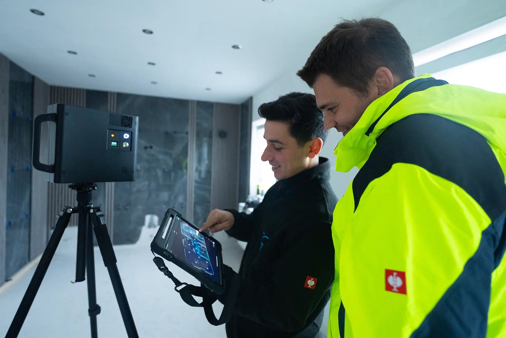 Zwei Personen in einem Raum, eine Person zeigt auf ein Tablet, das mit einem Gerät auf einem Stativ kabellos verbunden ist. Die Person rechts trägt eine leuchtend gelbe Jacke.