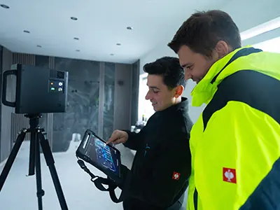 Zwei Personen in einem Raum, eine Person zeigt auf ein Tablet, das mit einem Gerät auf einem Stativ kabellos verbunden ist. Die Person rechts trägt eine leuchtend gelbe Jacke.