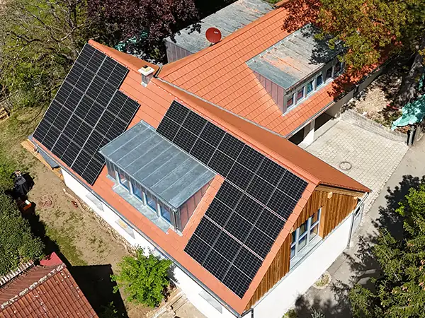 Vogelperspektive des sanierten Hauses. Auf dem Dach mit roten Ziegeln sind flächendeckend Solarzellen angebracht. Um das Gebäude sind die Zufahrt, Gärten und viele Bäume zu erkennen.