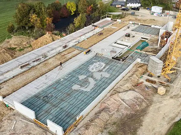 Luftaufnahme einer Baustelle mit Betonfundamenten, Baustellenkran und Erdarbeiten, umgeben von Bäumen und Feldern.