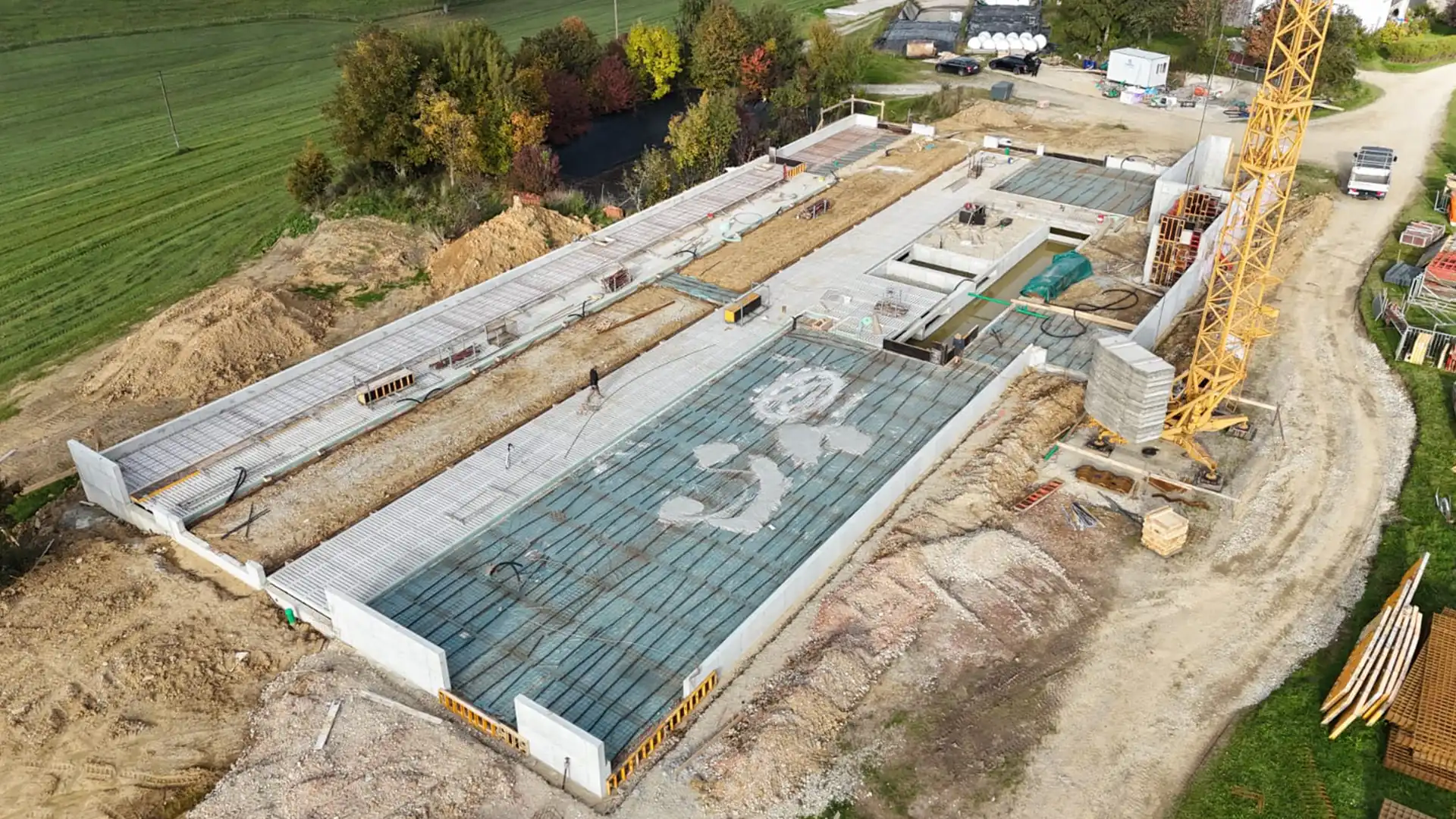 Luftaufnahme einer Baustelle mit Betonfundamenten, Baustellenkran und Erdarbeiten, umgeben von Bäumen und Feldern.
