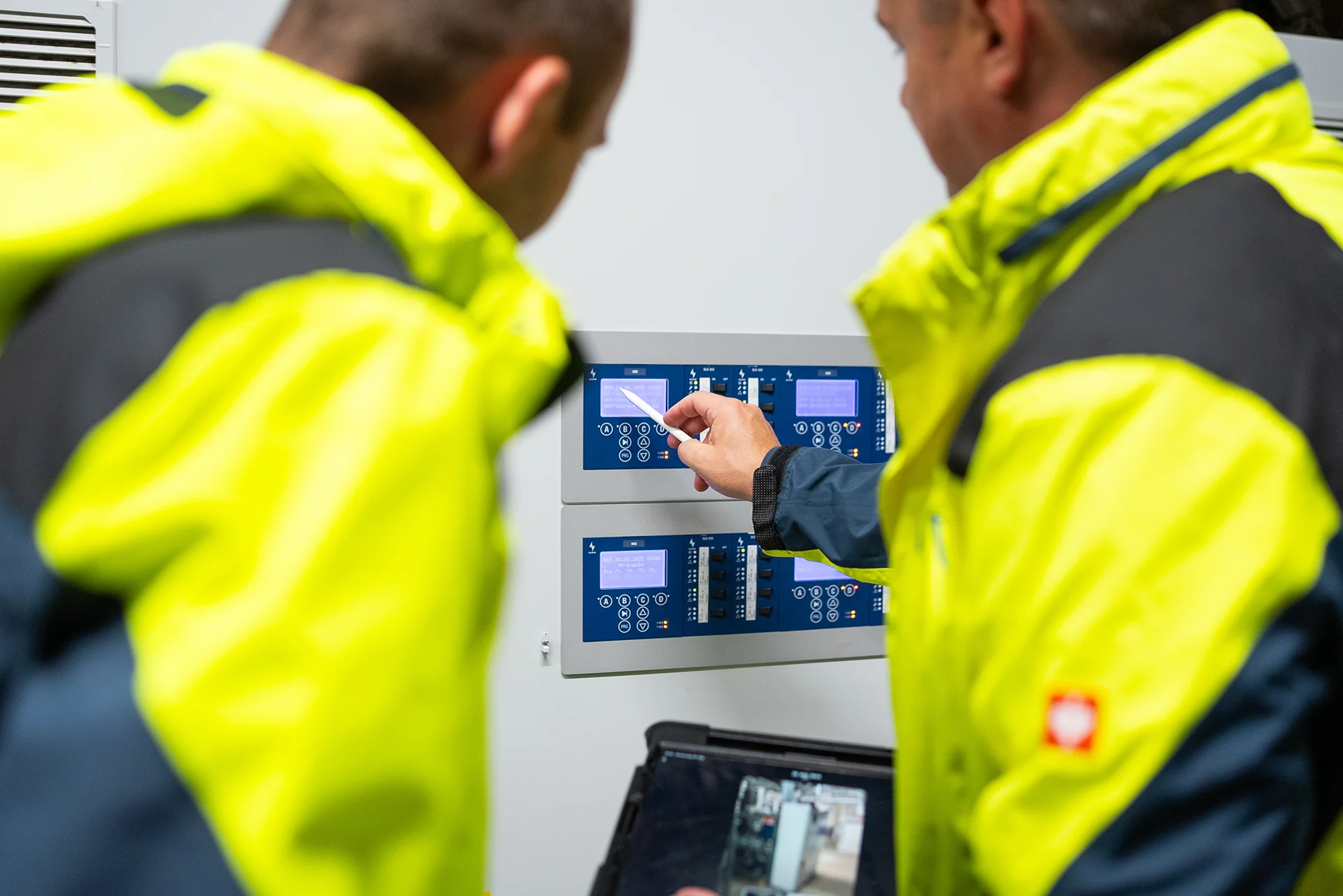 Zwei Personen in gelb-schwarzen Sicherheitsjacken schauen auf ein blaues elektronisches Bedienfeld mit Displays und Tasten. Die rechte Person hält ein Tablet mit 3D Scans in einer Hand, mit der anderen Hand hält sie einen Stylus auf das Bedienfeld.