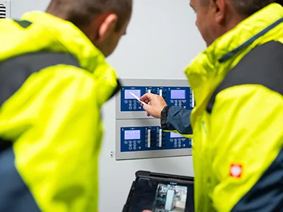 Zwei Personen in gelb-schwarzen Sicherheitsjacken schauen auf ein blaues elektronisches Bedienfeld mit Displays und Tasten. Die rechte Person hält ein Tablet mit 3D Scans in einer Hand, mit der anderen Hand hält sie einen Stylus auf das Bedienfeld.