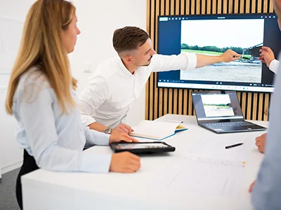 Vier Personen stehen an einem Tisch, eine zeigt auf einen großen Bildschirm mit dem Foto einer Baustelle, ein Laptop und ein Tablet liegen auf dem Tisch.