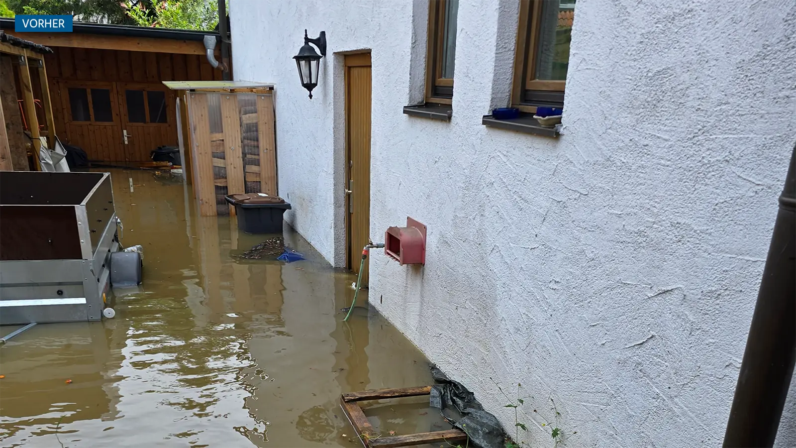 Haus während des Hochwassers. Das Wasser steht knöchelhoch um die Haustüre des Erdgeschosses. Das Wasser ist dunkel und matschig, das Haus ist durch das Hochwasser leicht verschmutzt.