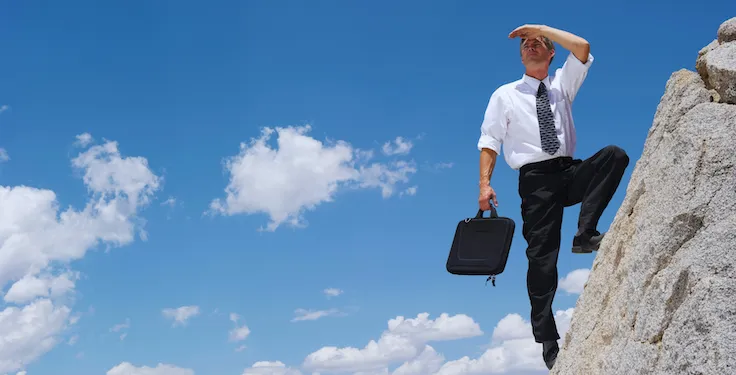 Businessman climbing a rock cliff while holding a briefcase and shading his eyes, looking into the distance under a blue sky with clouds.