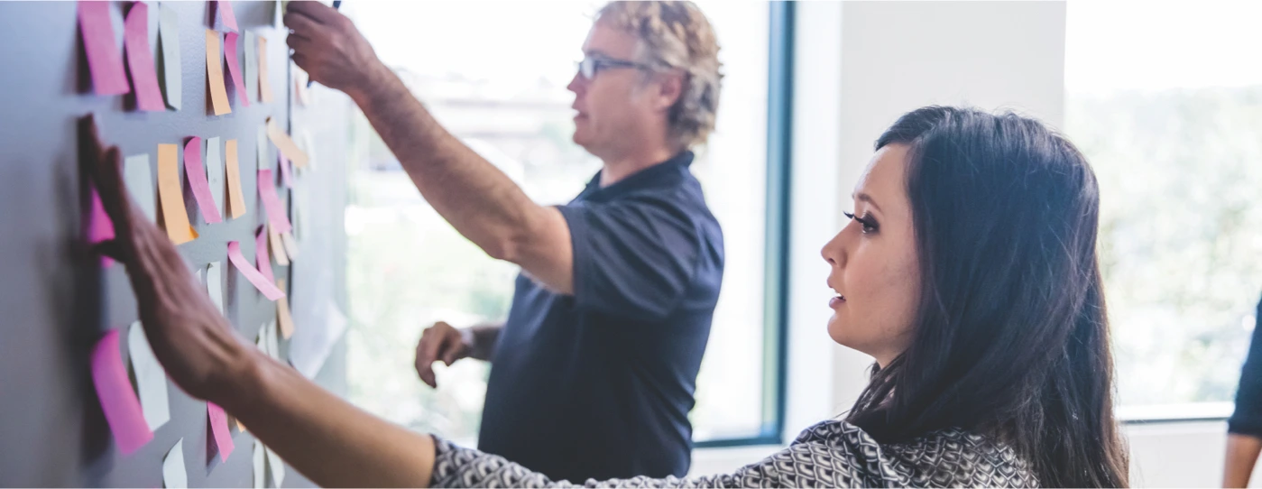 Two colleagues placing and organizing colorful sticky notes on a wall in a bright office.