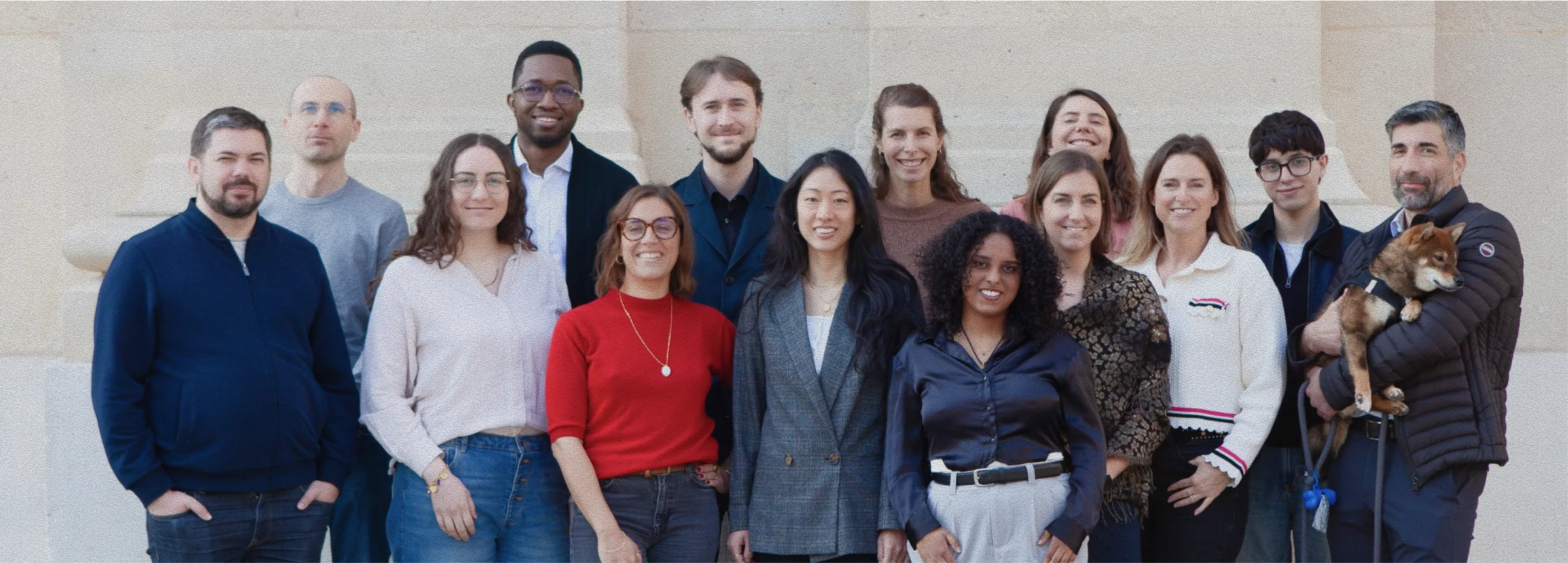 Group photo of fifteen people standing and smiling in front of a beige wall, one person holding a small brown dog.