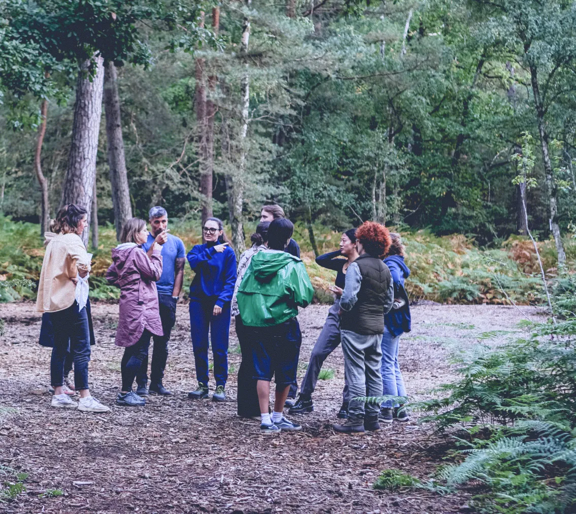 Group of people standing in a circle having a discussion in a forest clearing surrounded by trees and greenery.