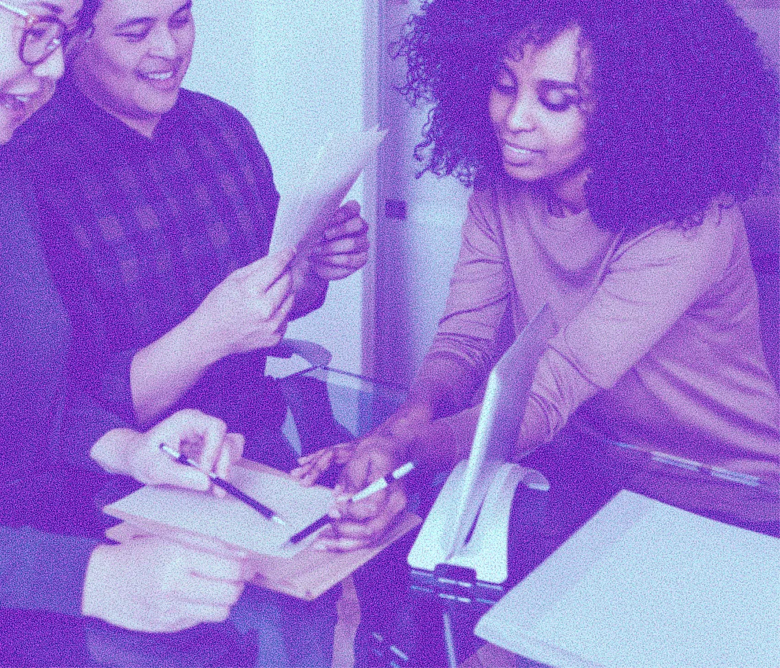 Three people discussing documents and writing notes at a glass table in a collaborative work setting.