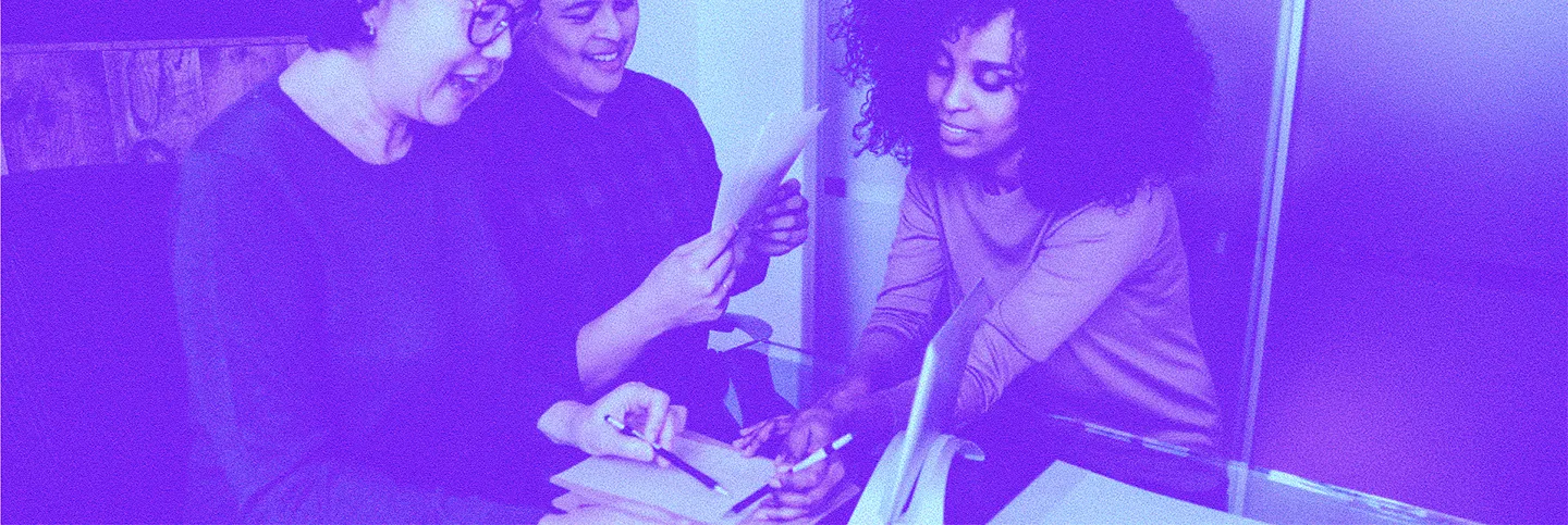 Three people engaged in a collaborative discussion, reviewing papers and taking notes at a desk.