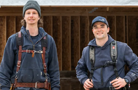 Two male construction workers wearing hoodies, harnesses, and hats standing inside a wooden building frame.