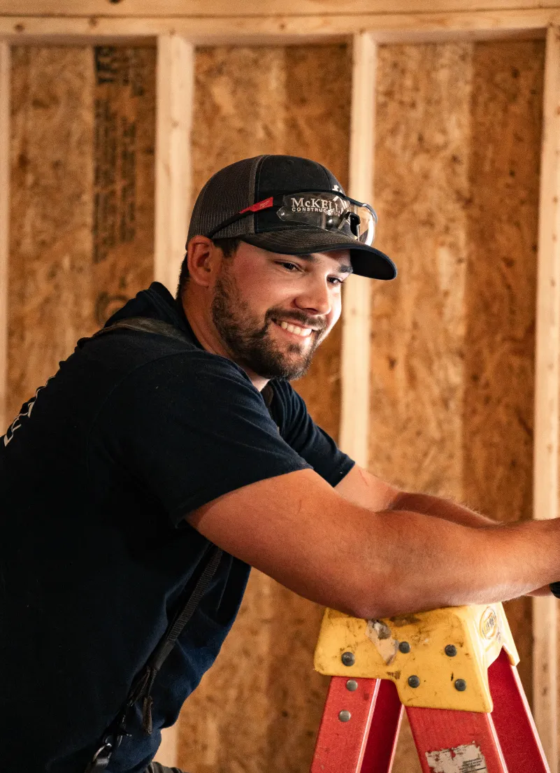 Smiling construction worker wearing a McKellar Construction cap and safety glasses leaning on a red and yellow ladder inside a wooden frame building.