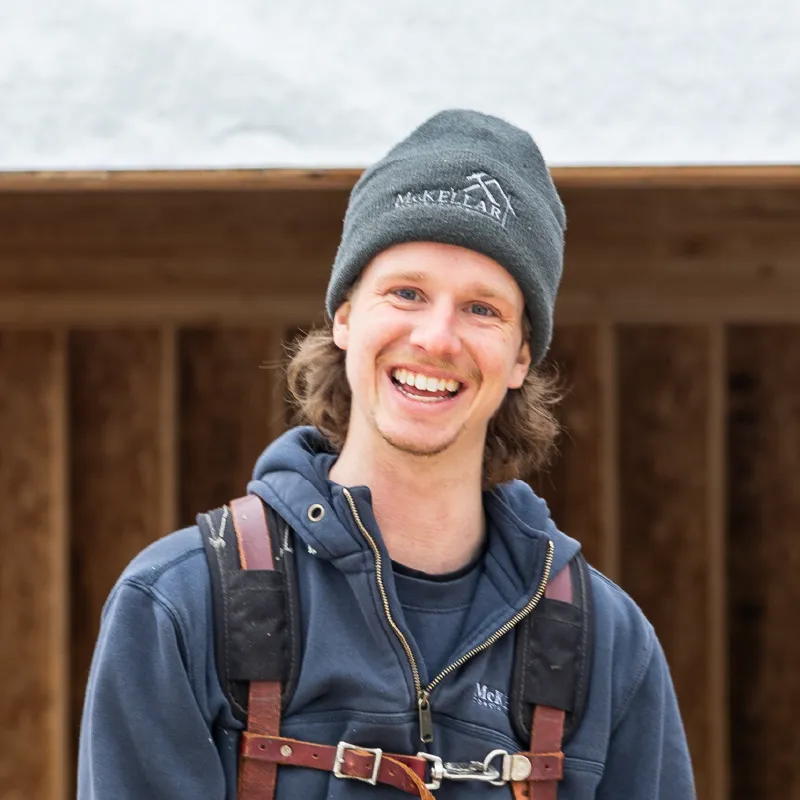 Smiling young man wearing a dark beanie and a hoodie with shoulder straps in front of wooden construction framing.