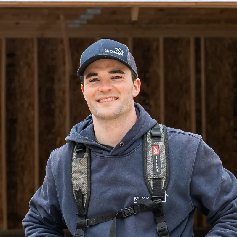 Young man wearing a navy cap and hoodie with harness straps, smiling in front of a wooden construction frame.