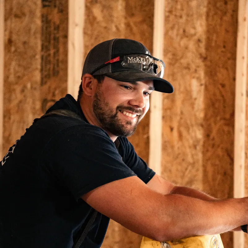 Smiling young male construction worker wearing a black McKEL construction cap and safety glasses, with wood framing background.