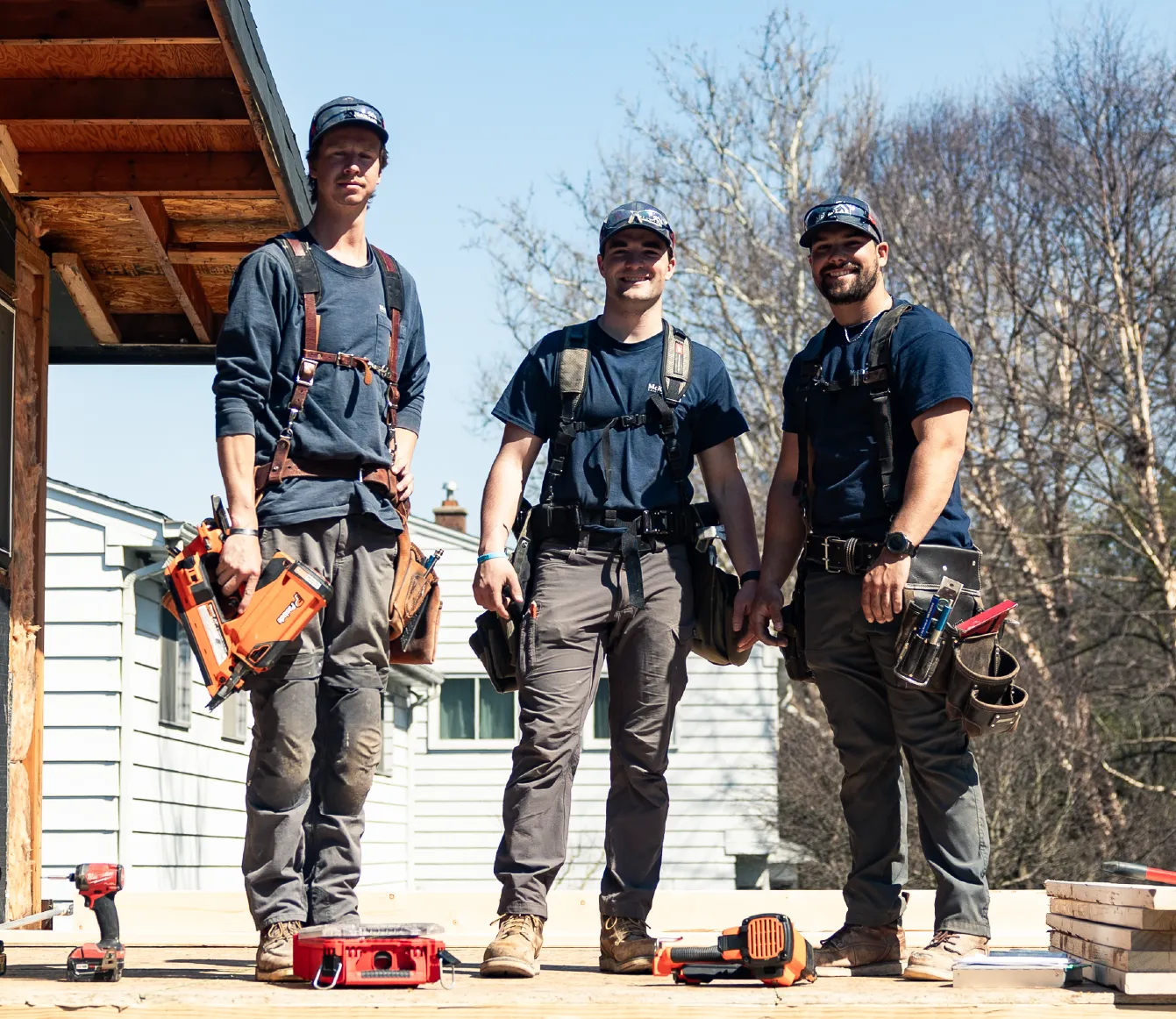 Three construction workers standing on a wooden deck with tools and equipment around them on a sunny day.