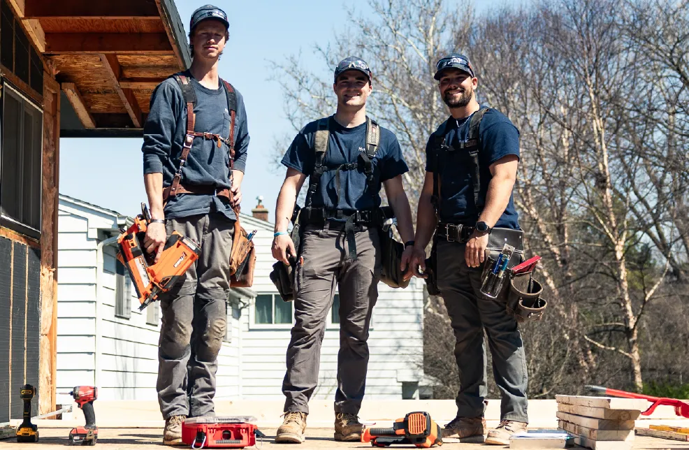 Three construction workers standing side by side on a wooden deck with tools and equipment around them.