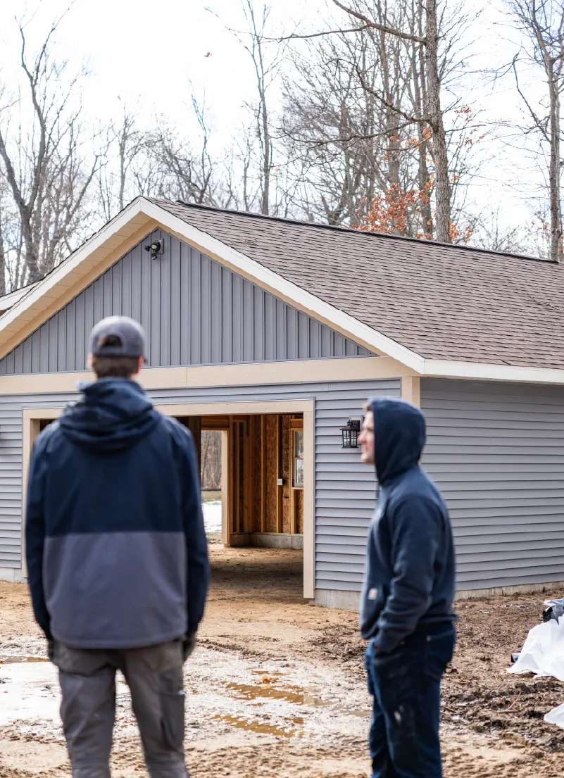 Two men in outdoor work clothes standing in front of a partially constructed gray barn with open entrance in a wooded area.
