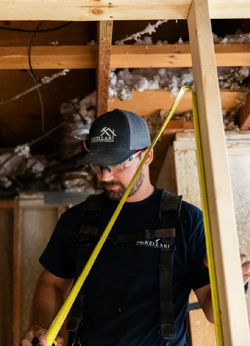 Construction worker wearing safety glasses and a cap measuring wood with a yellow tape measure inside a wooden framework.