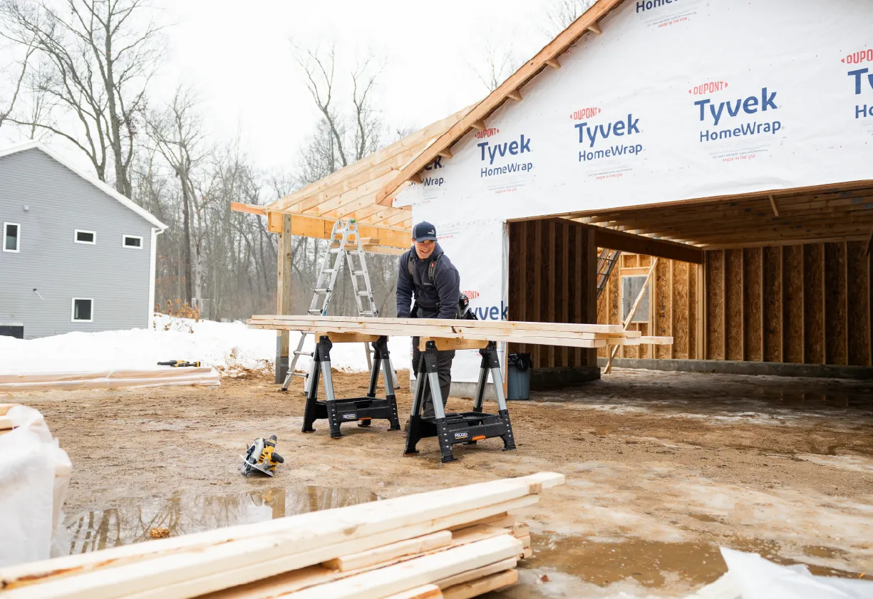 Construction worker cutting wooden boards on sawhorses in front of a house framing wrapped with Tyvek HomeWrap on a snowy day.