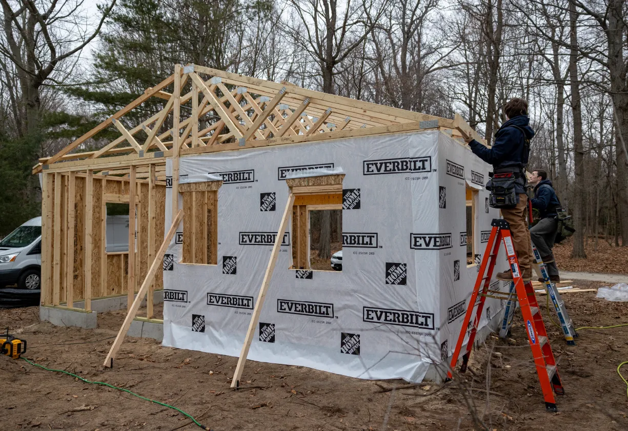 Two workers on ladders building a wooden house frame wrapped in Everbilt branded weather-resistant barrier in a forested area.