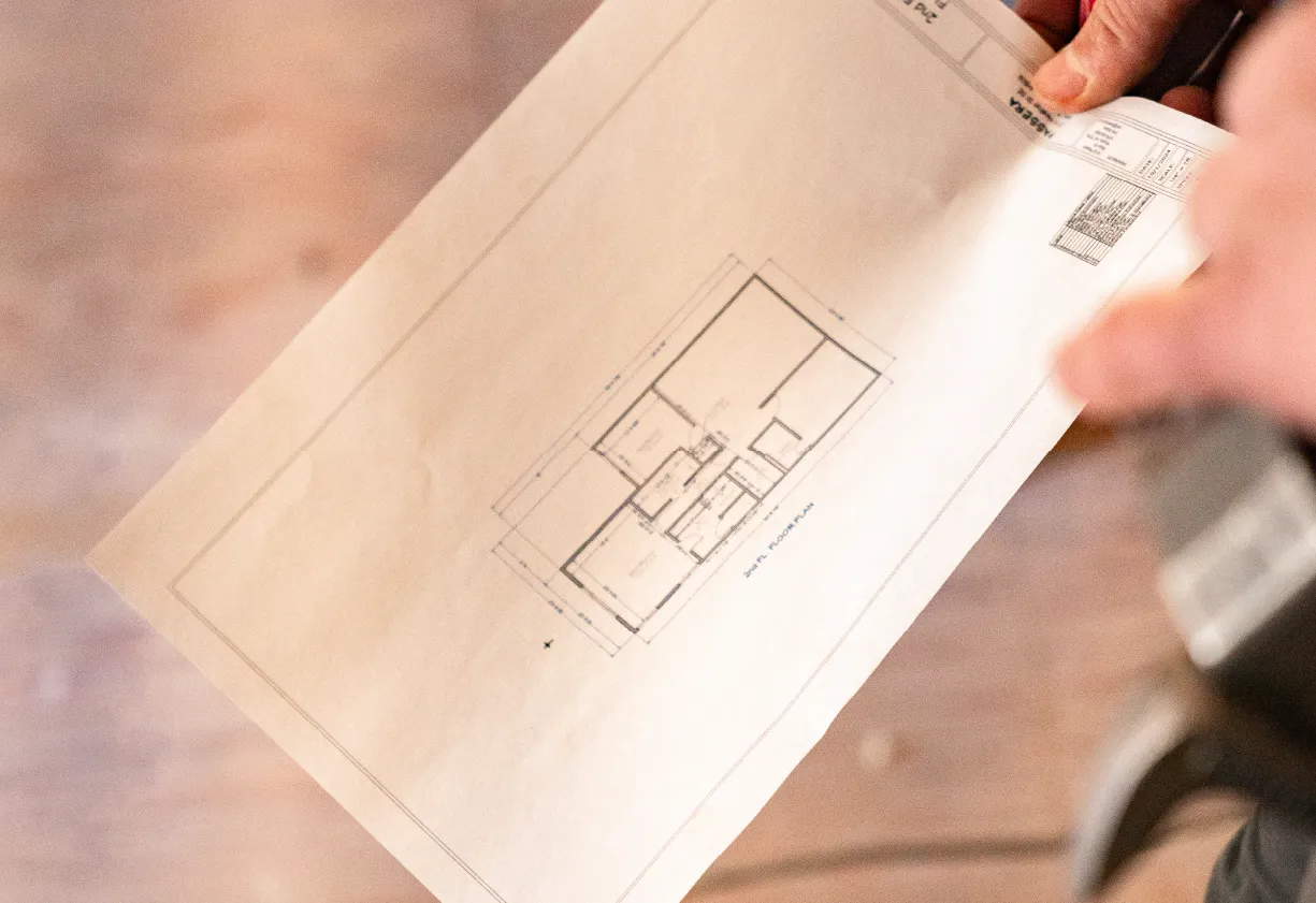 Hands holding a printed architectural floor plan drawing with room layouts and measurements.