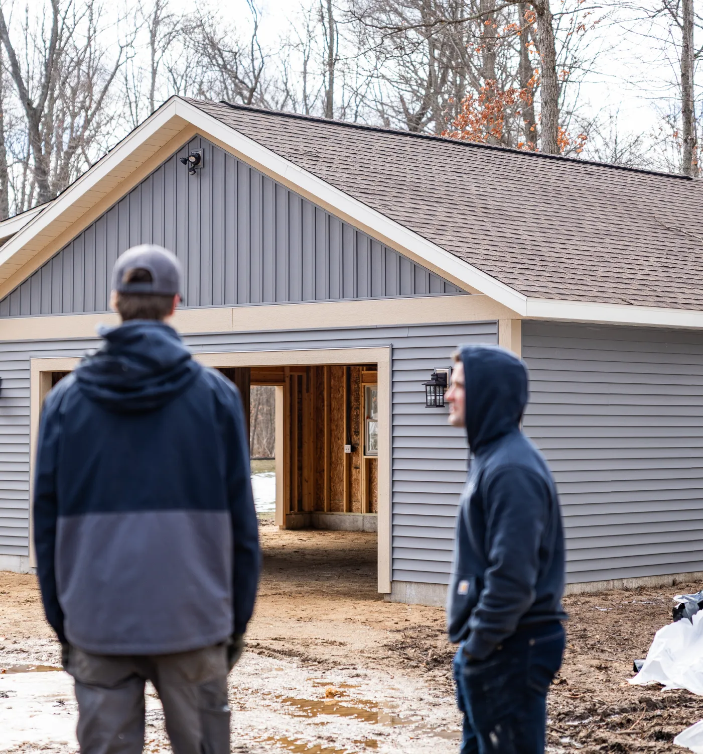 Two men in jackets standing outside a newly built gray garage with an open door showing unfinished interior walls.