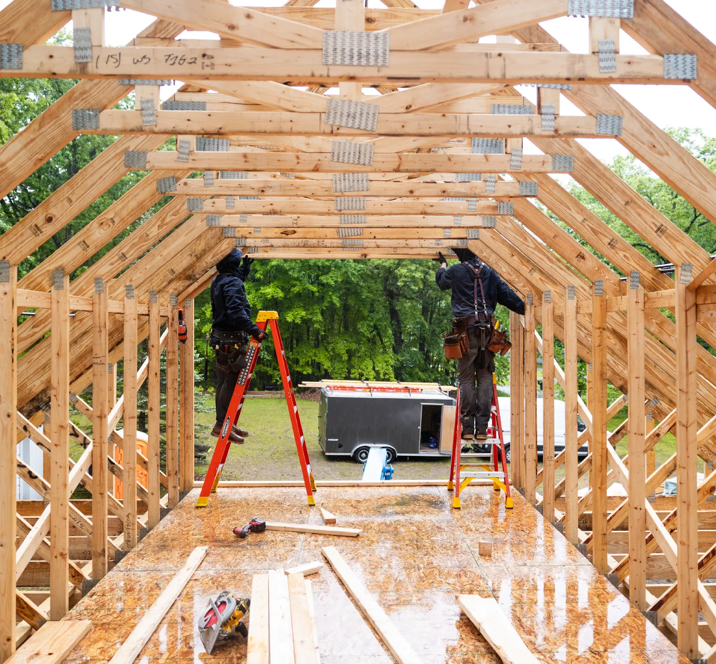 Two construction workers on ladders framing a wooden barn structure with tools scattered on the floor.