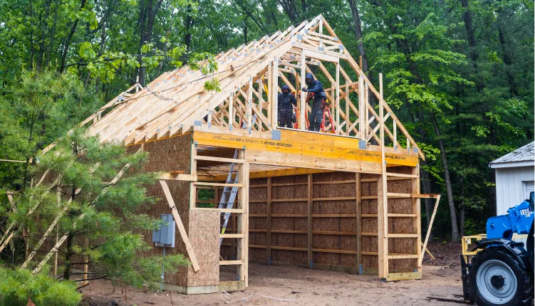 Wooden barn frame under construction in a forested area with two workers on scaffolding installing roof trusses.