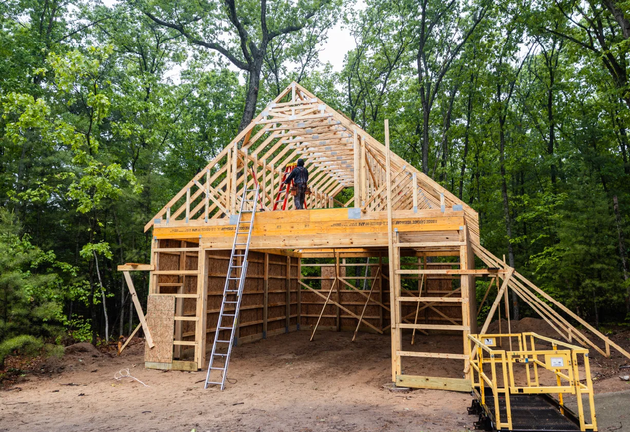 Wooden barn frame under construction in a forested area with a worker on a ladder installing roof trusses.
