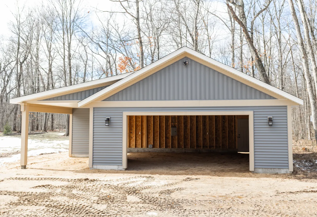 Newly built light blue garage with beige trim and an open garage door showing its unfinished wooden interior, surrounded by leafless trees.