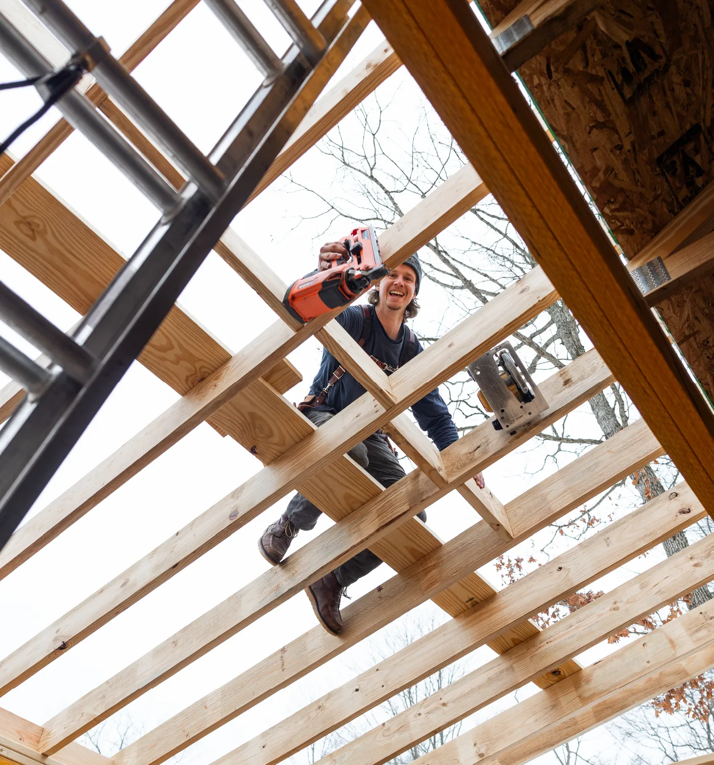 Smiling construction worker using a nail gun while standing on wooden beams of a building frame under construction.