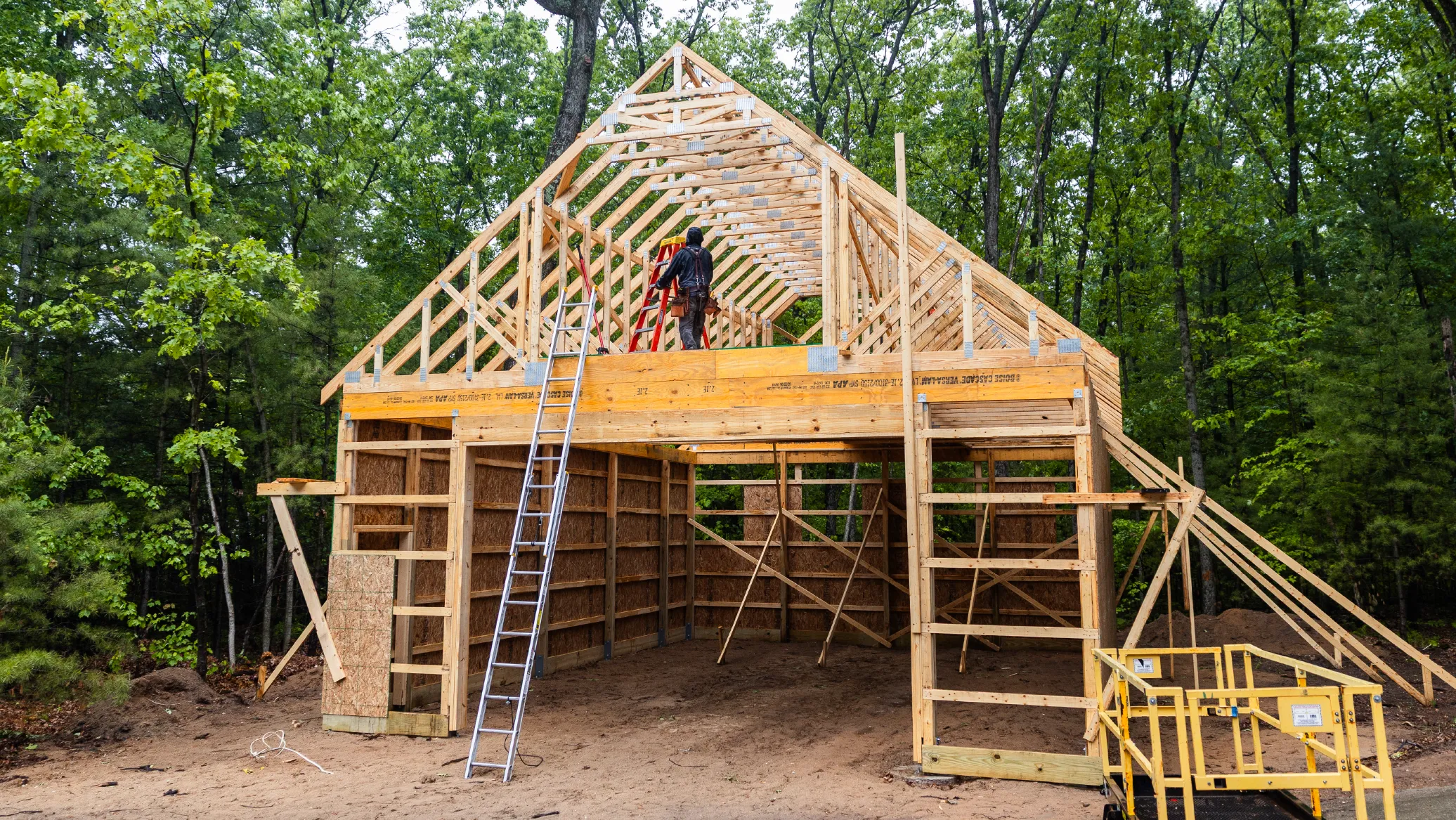 Construction worker on ladder framing wooden barn structure in wooded area.