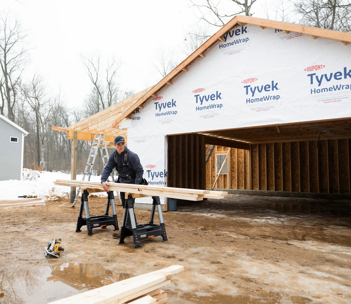 Construction worker handling wooden planks outside a partially built wooden structure wrapped in Tyvek HomeWrap in a snowy, wooded area.