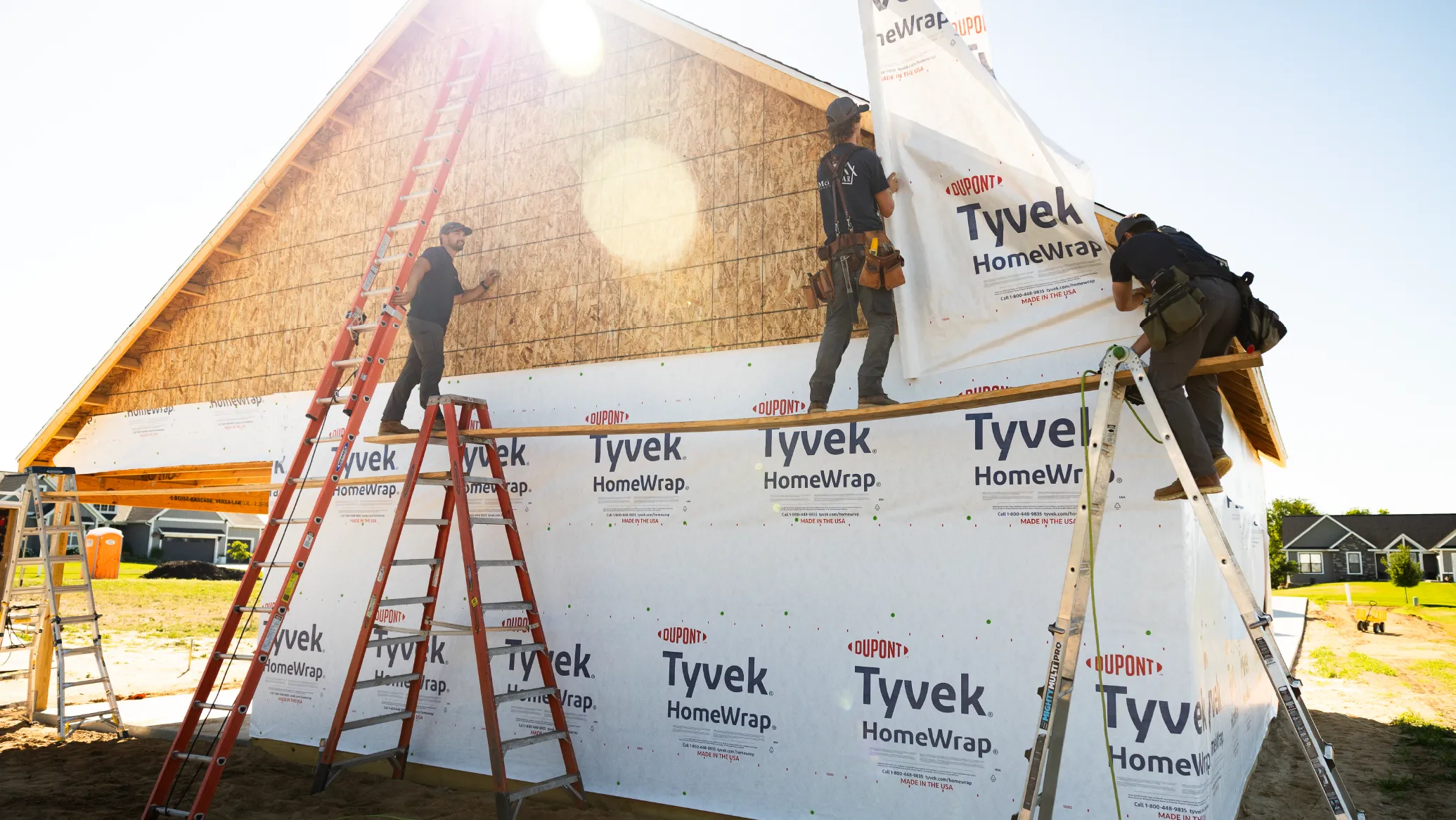Three construction workers installing Tyvek HomeWrap on the exterior of a wooden house frame using ladders and scaffolding.