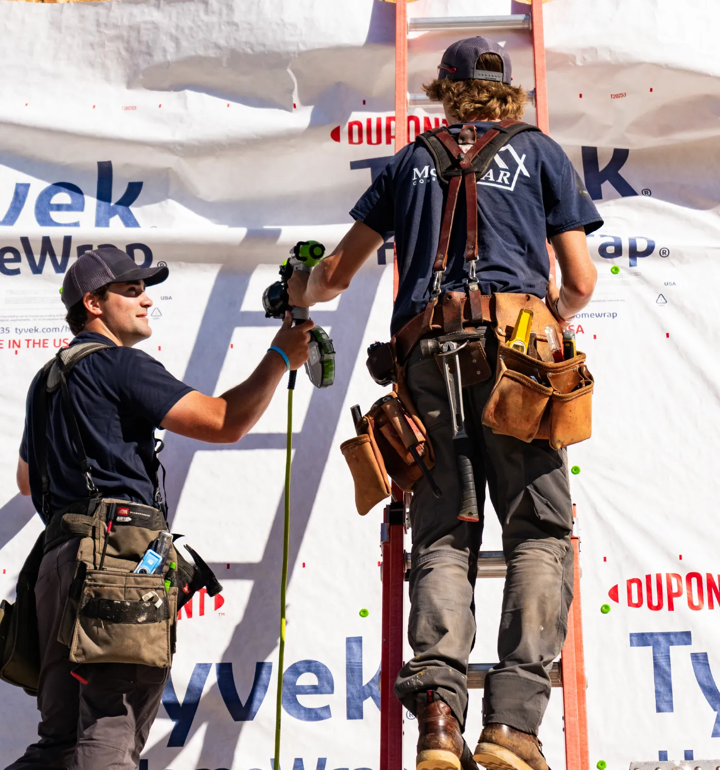 Two construction workers with tool belts working on a building covered in Tyvek home wrap, one on a ladder using a nail gun.