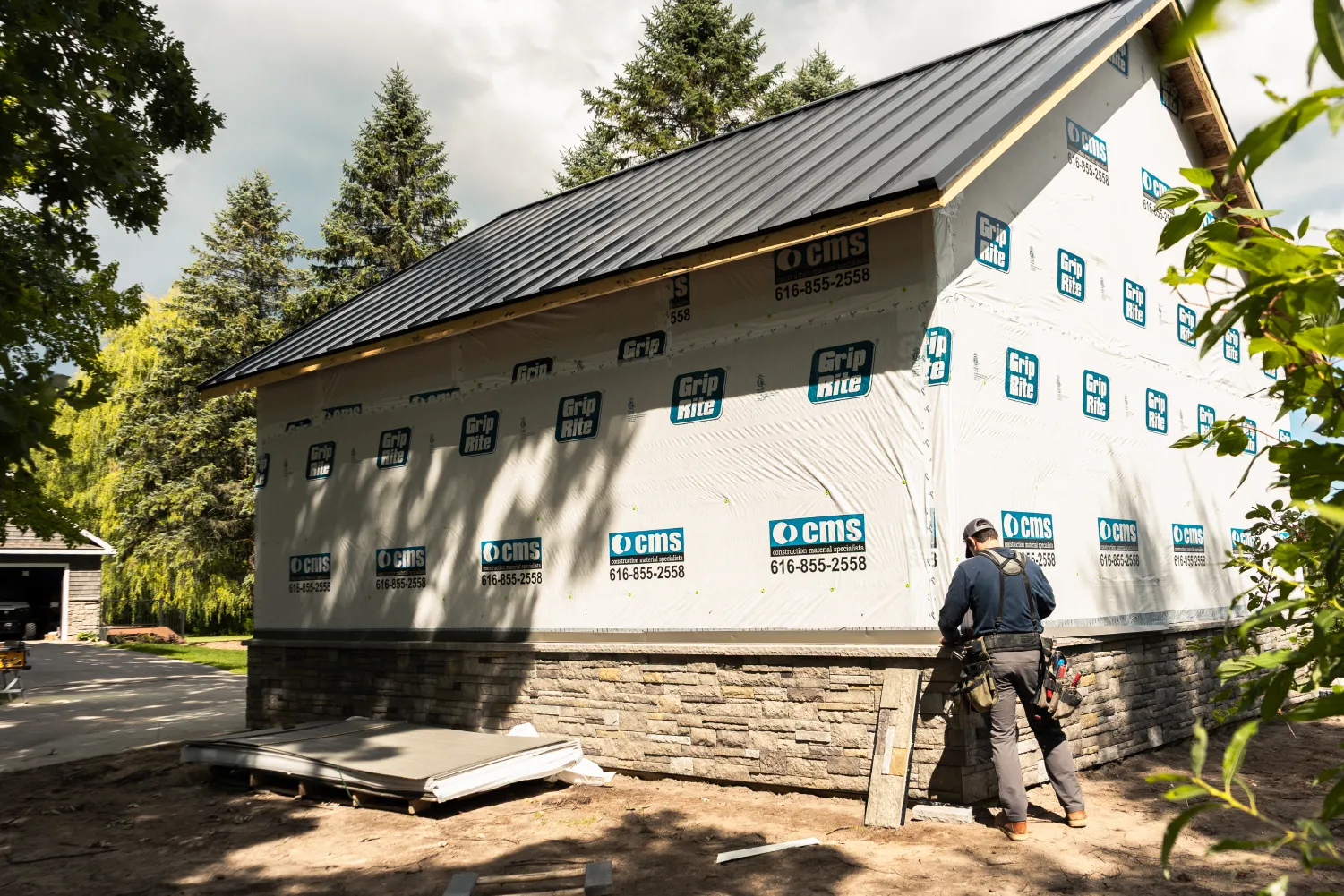Construction worker installing stone veneer on partially built house with black metal roof in a wooded area.
